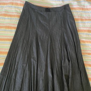 Grå långkjol från Birgitta - En elegant grå långkjol från Birgitta. Kjolen har en veckad design och är tillverkad av 65% viskos, 35% polyester. Den har en dragkedja och knappstängning i midjan.