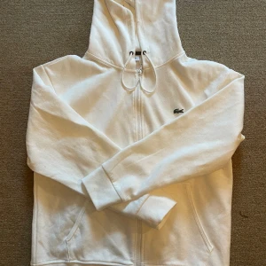 Lacoste Hoodie - En vit lacoste zip hoodie i storlek Large. Säljes då den inte passar mig
