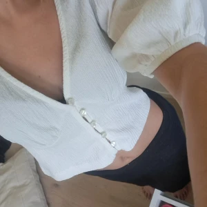 Vit blus med knappar - En vit blus med knappar framtill. Blusen har korta puffärmar och en v-ringad hals. Den är croppad och har en åtsittande passform.