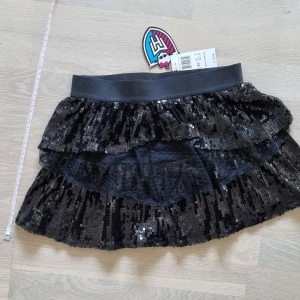 Oanvänd svart monster high kjol - Helt ny med prislapp på. Fodrad kjol från monster high med flera lager. Storlek M för barn 7-8 år. Måttband på bilderna finns som referens. Tappar dock lite glitter och några trådar sticker ut. Nypris: $16.99