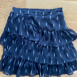 Svart kjol med blixtmönster från Sofie Schnoor - Säljer en superfin svart kjol från Sofie Schnoor med ett coolt blixtmönster. Kjolen har flera lager med volanger som ger en snygg look. Perfekt för både vardag och fest! Den är gjord av 100% polyester och är i storlek 152. Passar perfekt till en t-shirt eller en snygg blus. 🖤⚡