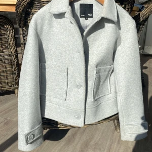 Ljusgrå ulljacka från 157 - Säljer en snygg ljusgrå kappa från lager 157. Jackan har en klassisk krage och stängs med knappar framtill. Den har två stora fickor på framsidan och är fodrad med ett glansigt material på insidan. Perfekt för höst och vår! 🧥