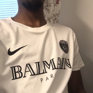 Vit fotbollströja från Nike och Balmain - Säljer en snygg vit fotbollströja från Nike i samarbete med Balmain. Tröjan har en stilren design med Balmain Paris-loggan tryckt på bröstet och PSG-emblemet på vänster sida. Perfekt för både träning och vardagsbruk. Tröjan är i nyskick och har korta ärmar.