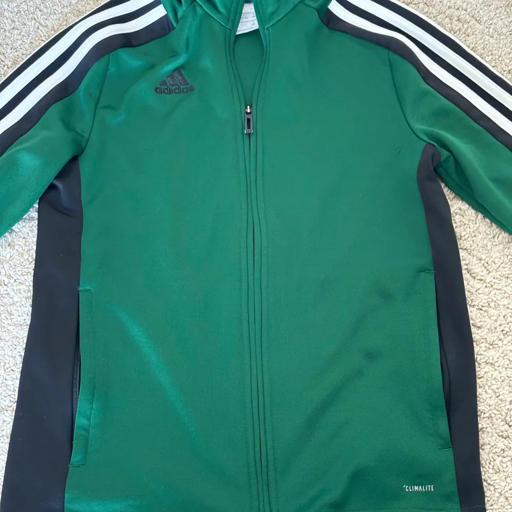 Säljer en grön träningsjacka från Adidas med svarta och vita detaljer. Jackan har dragkedja framtill och är tillverkad i polyester. Den har Adidas klassiska tre ränder längs ärmarna och är märkt med 'Climalite' för bra fukttransport. Perfekt för träning eller vardagsbruk.. Tröjor & Koftor.