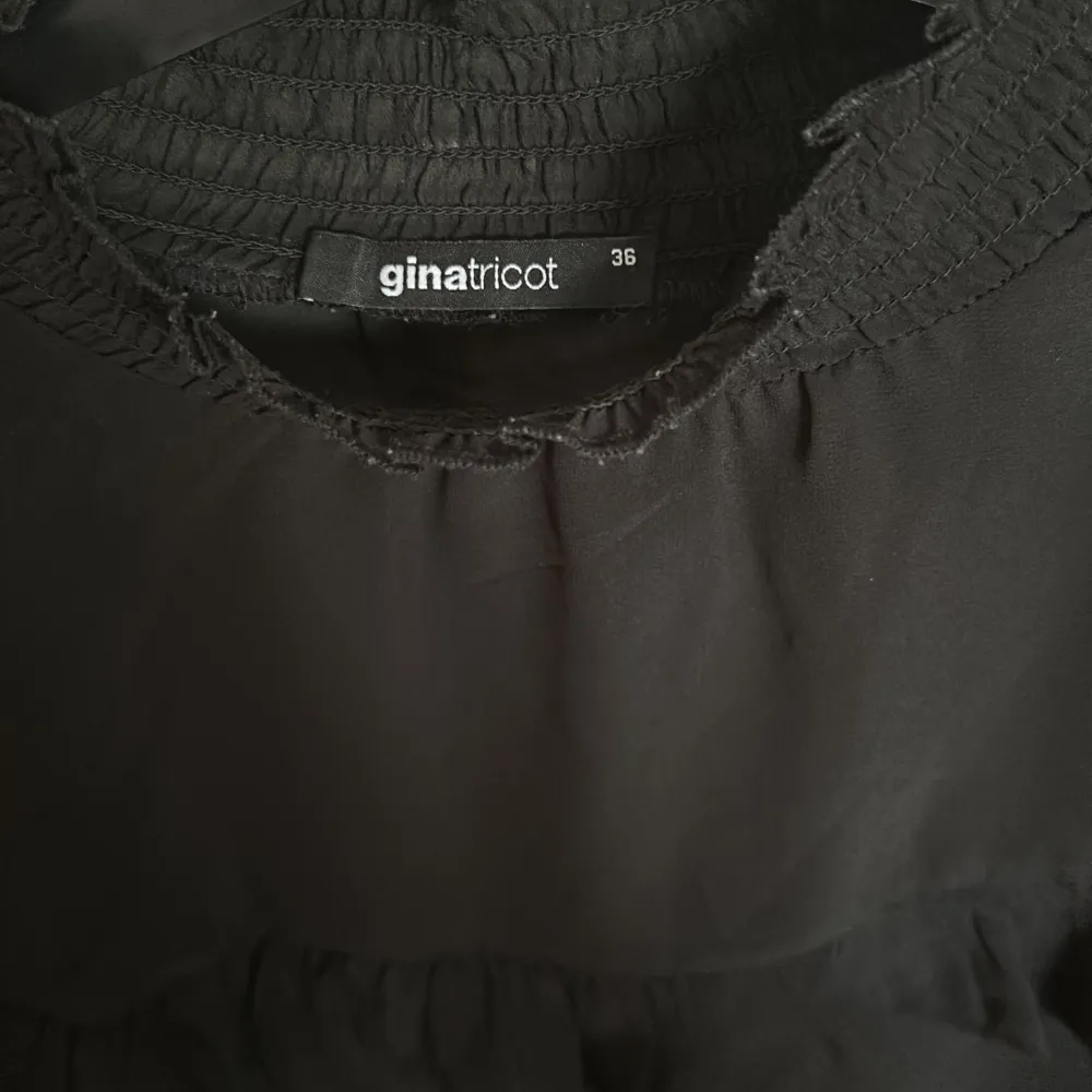 Säljer en elegant svart blus från Gina Tricot i storlek 36. Blusen har långa ärmar med volangdetaljer och en smockad halsringning samt ärmslut. Materialet är lätt och genomskinligt, perfekt för både vardag och fest. Blusen har en lös passform och är superbekväm att bära.. Toppar.