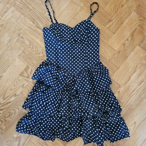 Mörkblå prickig klänning - En mörkblå kortklänning med vita prickar. Klänningen har smala axelband och volanger på kjolen. Den är ärmlös och har en åtsittande överdel med resår.