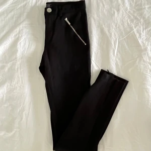 Svarta byxor från Zara - Svarta byxor från Zara med dragkedjedetaljer på framsidan samt längst ner. Tajta men stretchiga! Midjemått rakt över: 31.5cm. Innerbenslängd: 69cm. Nyskick.