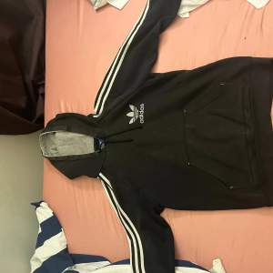 Säljer en svart hoodie från Adidas med vita ränder längs ärmarna och Adidas-loggan på bröstet. Den har en stor ficka på magen och en grå insida på huvan. Perfekt för höst och vinter! Har också en svart långärmad tröja med texten 'RAISE YOUR GAME' i stora vita bokstäver på framsidan. Båda är i bra skick och superbekväma.