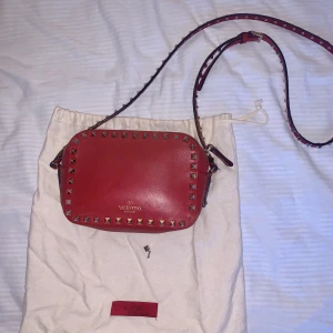 Valentino väska - Säljer min röda valentino rockstud väska. Den har använts flitigt och därav lite defekter. En nit har ramlat av (se bild 1) men jag skickar med extra nit så den går att lämna in och laga. Även den ljusa insidan har missfärgats lite. ❤️ Nypris ca 10.000kr