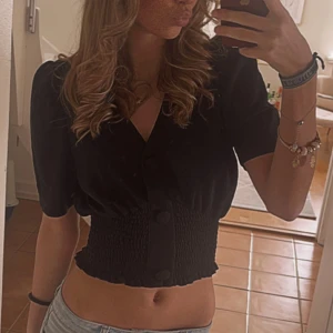 Svart blus från Stradivarius - En elegant svart blus från gina tricot med korta ärmar och v-ringning. Blusen har en smockad midja som ger en figurnära passform och knappar framtill.