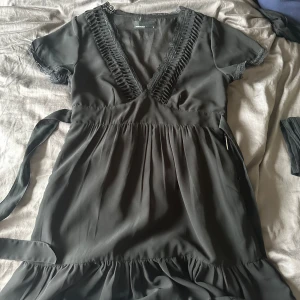 Svart klänning från Vero Moda - En elegant svart klänning från Vero Moda med v-ringning och spetsdetaljer. Klänningen har korta ärmar och ett knytband i midjan. Den är tillverkad i ett lätt och flödande material.