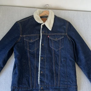 Blå jeansjacka från Levi's med foder - Aldrig använd blå jeansjacka från Levi's med vit foder i teddy-material. Jackan har knappar framtill och två bröstfickor med knappar. Den är fodrad med ett mjukt och varmt material, vilket gör den perfekt för kyligare väder.
