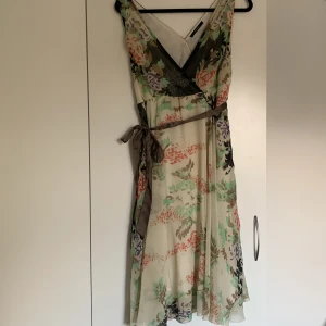 Blommig klänning från Vero Moda - En vacker blommig klänning från Vero Moda med ett knytband i midjan. Klänningen har en v-ringad hals och är ärmlös. Den har en lätt och luftig design med ett blommigt mönster i olika färger på en ljus bakgrund.