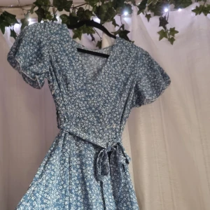 Blommig klänning  - Blå och vit blommig klänning, sydd som en omlottsklänning, med snöre runtom. Liten volang nertill samt en dragkedja på sidan. Strl. S men passar också för XS 