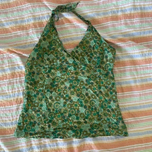 Grön blommig halterneck topp från Indiska - En grön blommig halterneck topp från Indiska i storlek M. Toppen är ärmlös och har ett knytband runt halsen. Den är tillverkad av 100% bomull och har ett färgglatt blommönster i gröna och gula nyanser.