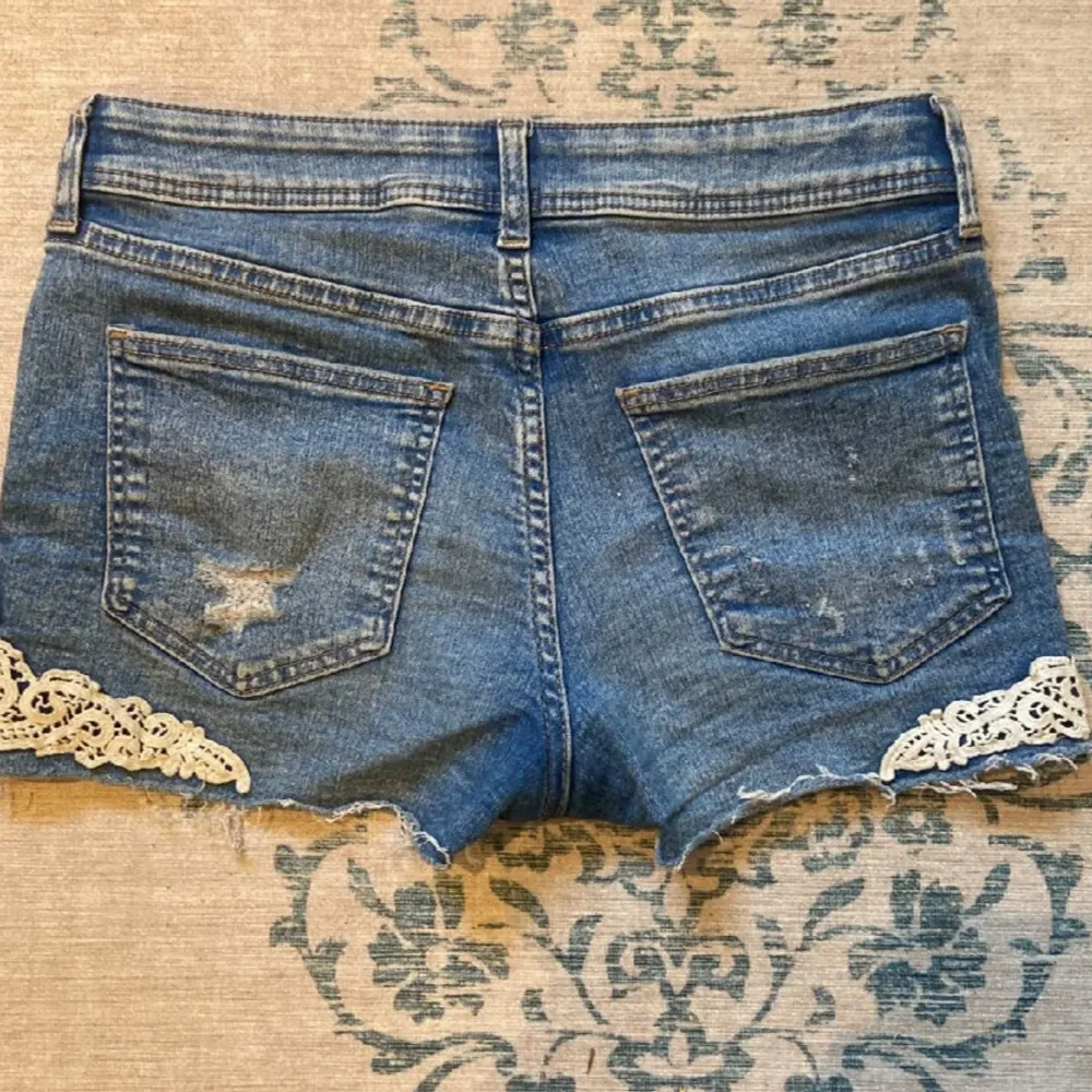 Säljer dess super snygga blå jeansshorts från H&M med slitna detaljer och spetsapplikationer på sidorna. Shortsen har en låg midja och fransiga kanter.. Shorts.