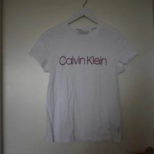 Calvin Klein T-shirt vit - Vit t-shirt med rosa Calvin Klein-logotyp. T-shirten är i gott skick använd ca 1 gång  Storlek L men liten i passformen  Material: Bomull Märke: Calvin Klein 