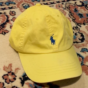 Gul Ralph Lauren keps med blå broderad logga - Gul justerbar Polo Ralph Lauren keps med mörkblå broderad logga + märkets namn ”POLO” på baksidan 🧢  Så snygg till en outfit som redan har gula och/eller mörkblåa element men också som pricken över i:et till en outfit som behöver den extra perfekta accessoaren!  Alla priser är inkl. frakt och alla plagg tvättas innan de skickas iväg