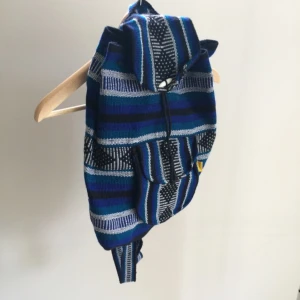  - Mönstrad rymlig ryggsäck i blått, vitt och svart som jag tror är handgjord. Ryggsäcken har ett stort fack som stängs med en oval vit plast "knapp". Det finns ett snöre att dra åt öppningen med. 
På nedre framsidan finns ett mindre fack som stängs med karborre. 
De två axelremmarna går ej att reglera och är ganska långa. 
Finns en mindre ögla högst upp på ryggan. 