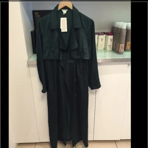  - En super snygg trenchcoat i satin. Brett, avtagbart knytskärp i midjan samt sidfickor. Höga slitsar i sidorna.  Säljer pga fel storlek. Aldrig använd. Ord. Pris 699:-