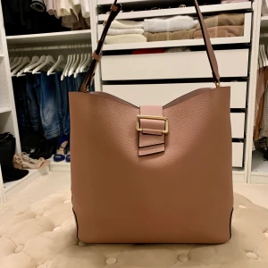 Hoboväska i nude/rosa - Hobo väska i finaste nude/rosa färgen i nyskick. Använd 2 gånger. Väskan har en tillhörande clutch med innerfack som ingår samt ett långt axelband. Mycket rymlig - får plats med en dator. Mått: 34x31 cm. Djup: cirka 13 cm. Köparen står för frakt.