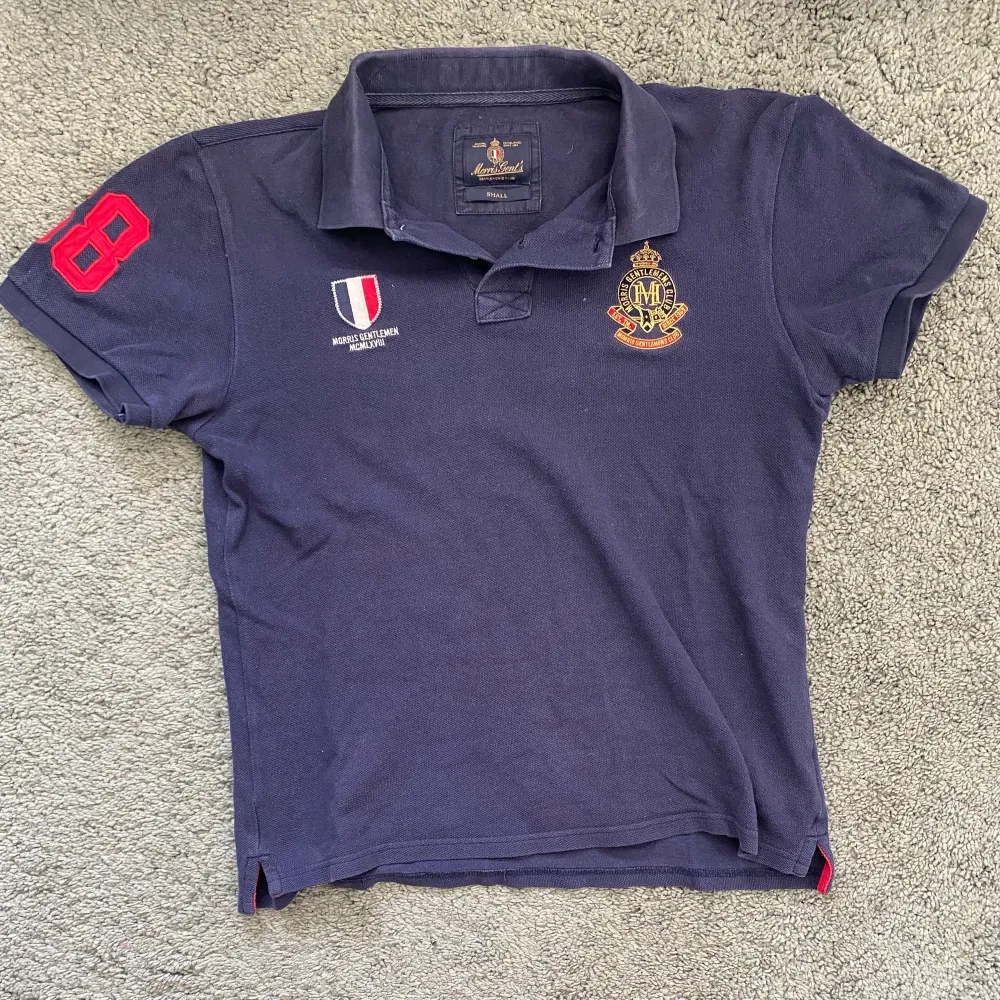 En marinblå Morris piké med ”68” på högra ärmen och två emblem på bröstet. Bra skick. Storlek Xs. T-shirts.