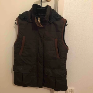  - Väst från JH collection (hööks), med avtagbar luva och lite ludd i kragen. Varm och skön, sitter snyggt, bra fickor som går att knäppa. Mörkbrun med guldig dragkedja och ljusbruna manchester detaljer.