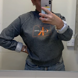 Grå Calvin Klein tröja - En grå Calvin Klein tröja med orange tryck, använd typ 2 gånger så den är nästan i nyskick, jätte fin men har inte haft användning för den, köp på Jolina i Borås city för 1200kr