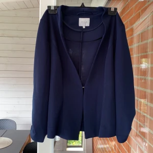 Kavaj  - En figursydd kavaj från Vila i marinblå med ett litet knäppe i mitten samt med uppsydda ärmar. Kavajen är i fint skick. Frakten är inräknad i priset.