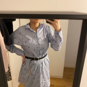 Blå Skjortklänning med blommor - En sommrig skjortklänning som är lite stor i storleken. Storlek S men som sagt, den är lite stor