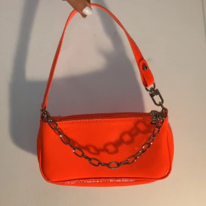 Axelväska - Liten neon-orange färgad baguette stil handväska. Lackat material med silver detaljer. Handtag avtagbar på ena sidan. Helt oanvänd. Köparen står för frakt ifall man vill ha det fraktat. 