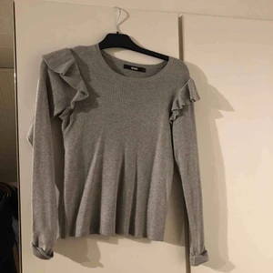  - Grå stickad tröja med volanger på axlarna.