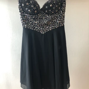 Festklänning - Svart festklänning med både nitar och strasstenar. Axelbandslös men med fästen för att sätta dit löstagbara axelband. Tight överdel med lös kjol. Frakt tillkommer om man inte kan möta upp i Växjö. 