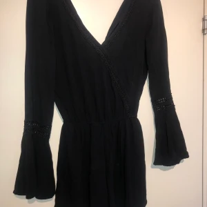 Svart playsuit, stl M - Svart trumpetärmad byxdress med knapp i ryggen och spets-liknande detaljer på ärmarna och v-ringningen. Fint skick och sparsamt använd. 