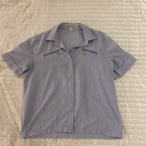 Blå-och vitrandig topp - Blå-och vitrandig topp med knappar och två framfickor med dragkedja. Strl S. OBS! Ett litet hål finns under sista knappen på vänster sida av toppen och tyvärr saknas en knapp - syns däremot inte när man bär toppen.