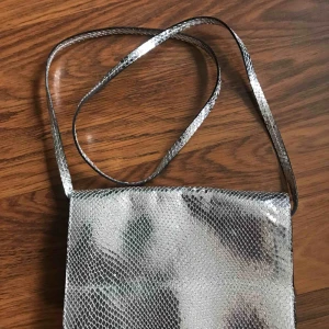  - Handväska i äkta skinn, i metallic med reptilmönster, är köpt på And other stories och endast använd ett fåtal gånger. I jättefint skick! skickar med postnord skicka lätt för 58kr.  Mått ca: Bredd 23,5 cm Höjd 15,5 cm Djup 5 cm Axelbandet 120 cm