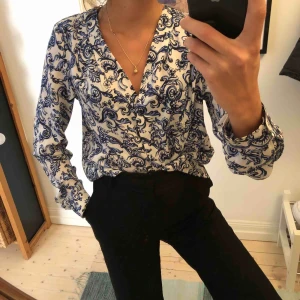  - Blus/skjorta med knappar i mönstrat tyg med urringning framtill. 