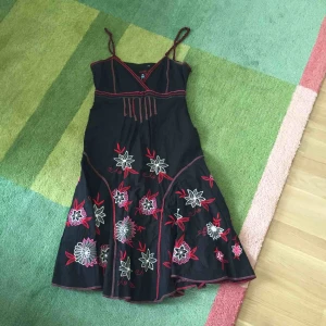  - Desigual fin klänning i svart med brodyr i rött, rosa, grått och vitt. Snurrat band runt ena axelbandet behöver fästas och deofläckar behöver tas bort (bild 2). Även skada i tyget i nedre delen av dragkedjan (bild 3).