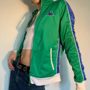 Vintage grön, blå & vit 80s Kappa tränings tracksuit jacka - Vintage Kappa tränings/tracksuit jackagrön i en klar grön färg med mörkblåa ränder längs utsidan av armarna och vid sidan av magen + smalare vita kontrasterande ränder vid sidan om de mörklblåa och vid fickorna för att få färgerna att poppa 🧩 Så snygg både till en mer retro 80s look men också perfekt för en modern outfit inspirerand av  the 2000s med ett par lågmidjade jeans och en vit topp under, eventuellt ett par färgglada solglasögon i matchande färger för att steppa up your game!
