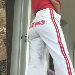 Skitcoola vita byxor - Vita relativt lågmidjade utsvängda byxor med röda detaljer. Röda linjer längs benen och röd text på rumpan. Använda Max två gånger men har några fläckar längst ner på baksidan