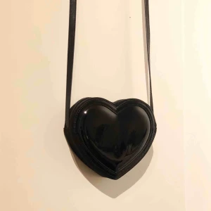 - Svart liten hjärtformad väska i ett lackat material på ena sidan, andra sidan är matt ”sammet” Facket är inte jättestort, men mobil och plånbok får plats! Bandet är inte reglerbart. Frakt tillkommer💞