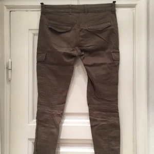  - Säljer ett par grön-bruna byxor med fickor på benen och dragkedjor längst ned. Från Lindex, storlek 38.