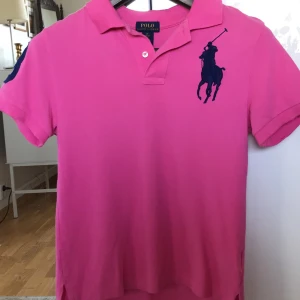 T-shirt, Ralph Lauren - Klassisk piké från Ralph Lauren i en nästan chockrosa färg och med marinblått motiv. Som i nyskick! Passar en XS