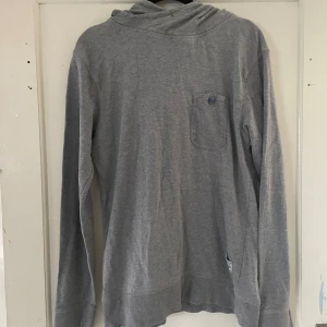 Grå hoodie från Jack & Jones - Säljer en grå hoodie från Jack & Jones, perfekt för en avslappnad stil. Den har en liten bröstficka med knapp och är tillverkad i ett mjukt material som känns skönt mot huden. Passar bra till både vardag och chillkvällar.