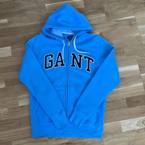 Blå hoodie från GANT - Säljer en snygg blå hoodie från GANT i storlek M. Den har en dragkedja framtill och stora bokstäver 'GANT' tryckt över bröstet. Perfekt för en avslappnad stil och passar bra till både höst och vår. Huvan har vita snören för justering.