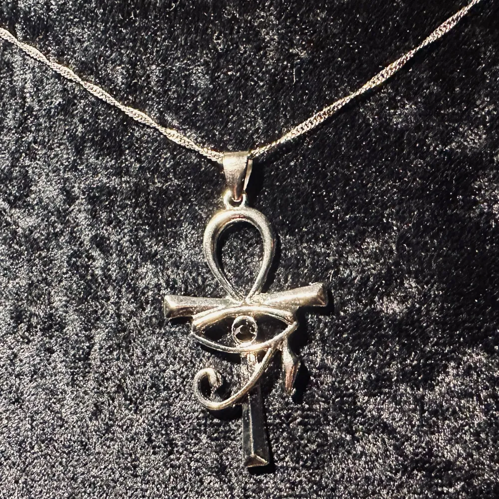 Nytt halsband i rostfritt stål med ankh-symbol och Ras öga med svart sten. Symboliserar liv, evigt liv och gudomligt beskydd. Kallas ibland vampyrkors. Stilrent och kraftfullt.    Amuletten är 4,5 cm lång x 2,7 cm bred. 45  + 5 cm kedja. Nytt.. Accessoarer.