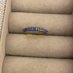 Guldring med blå stenar - Säljer en elegant guldring med en rad av små blå stenar. Ringen är gjorde av rostfritt stål och är guldpläterad. Den blå färgen på stenarna ger en fin kontrast mot det gyllene guldet. Perfekt som en subtil men vacker accesso