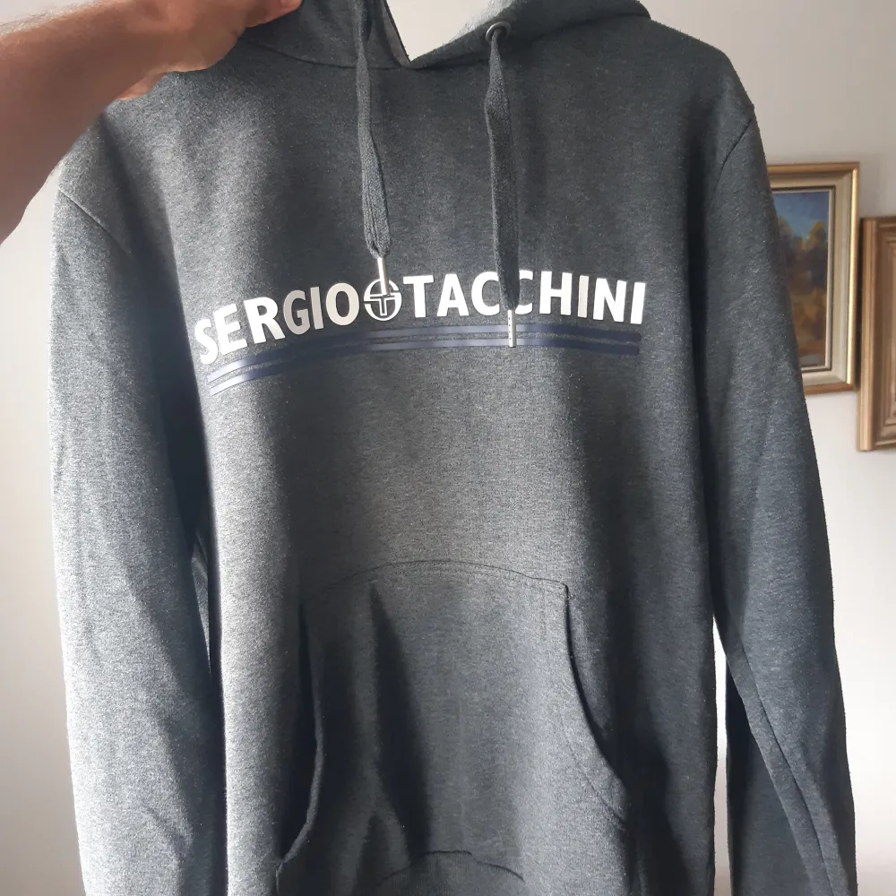 Säljer en snygg grå hoodie från Sergio Tacchini i storlek M. Den har en klassisk design med loggan tryckt i vitt och blått på bröstet. Hoodien är perfekt för både vardag och träning, med en bekväm passform och mjukt material. Den har en känguruficka framtill och justerbar huva med dragsko. Perfekt för höst och vinter!. Hoodies.