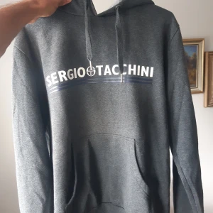 Grå hoodie från Sergio Tacchini - Säljer en snygg grå hoodie från Sergio Tacchini i storlek M. Den har en klassisk design med loggan tryckt i vitt och blått på bröstet. Hoodien är perfekt för både vardag och träning, med en bekväm passform och mjukt material. Den har en känguruficka framtill och justerbar huva med dragsko. Perfekt för höst och vinter!