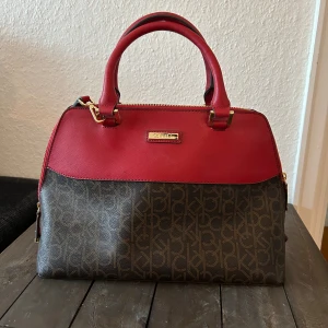 Röd och brun handväska med monogram - Säljer en snygg handväska från Calvin Klein i rött och brunt. Väskan har ett klassiskt monogrammönster och är gjord i syntetmaterial. Den har både handtag och en avtagbar axelrem, vilket gör den superpraktisk. Insidan är rymlig med flera fack och dragkedja för säker förvaring. Nyskick 